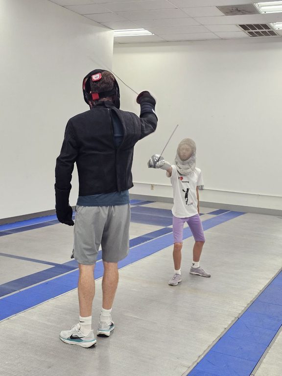 A coach giving an individual lesson to a young fencer in a mask on the strip