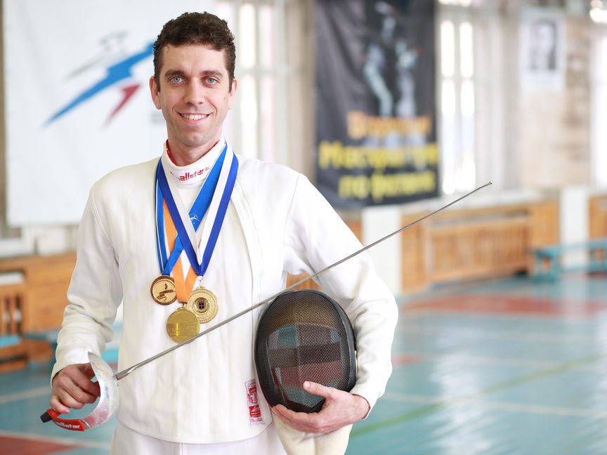 Coach Pavel Bykov in fencing whites with his medals, holding his mask and sabre