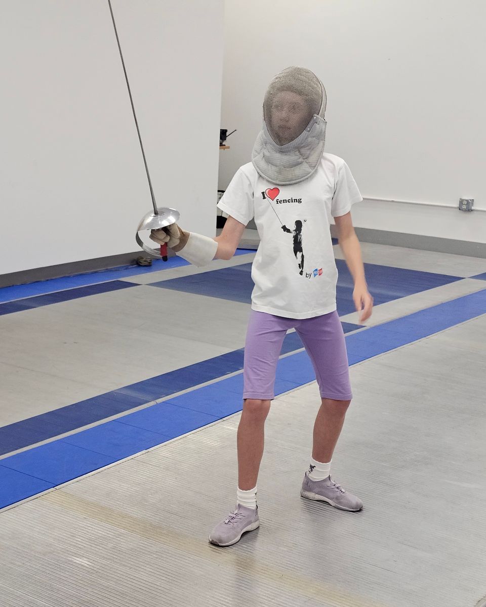 A young fencer in a mask and T-shirt holds the en garde stance on the strip at West Coast Fencing Academy