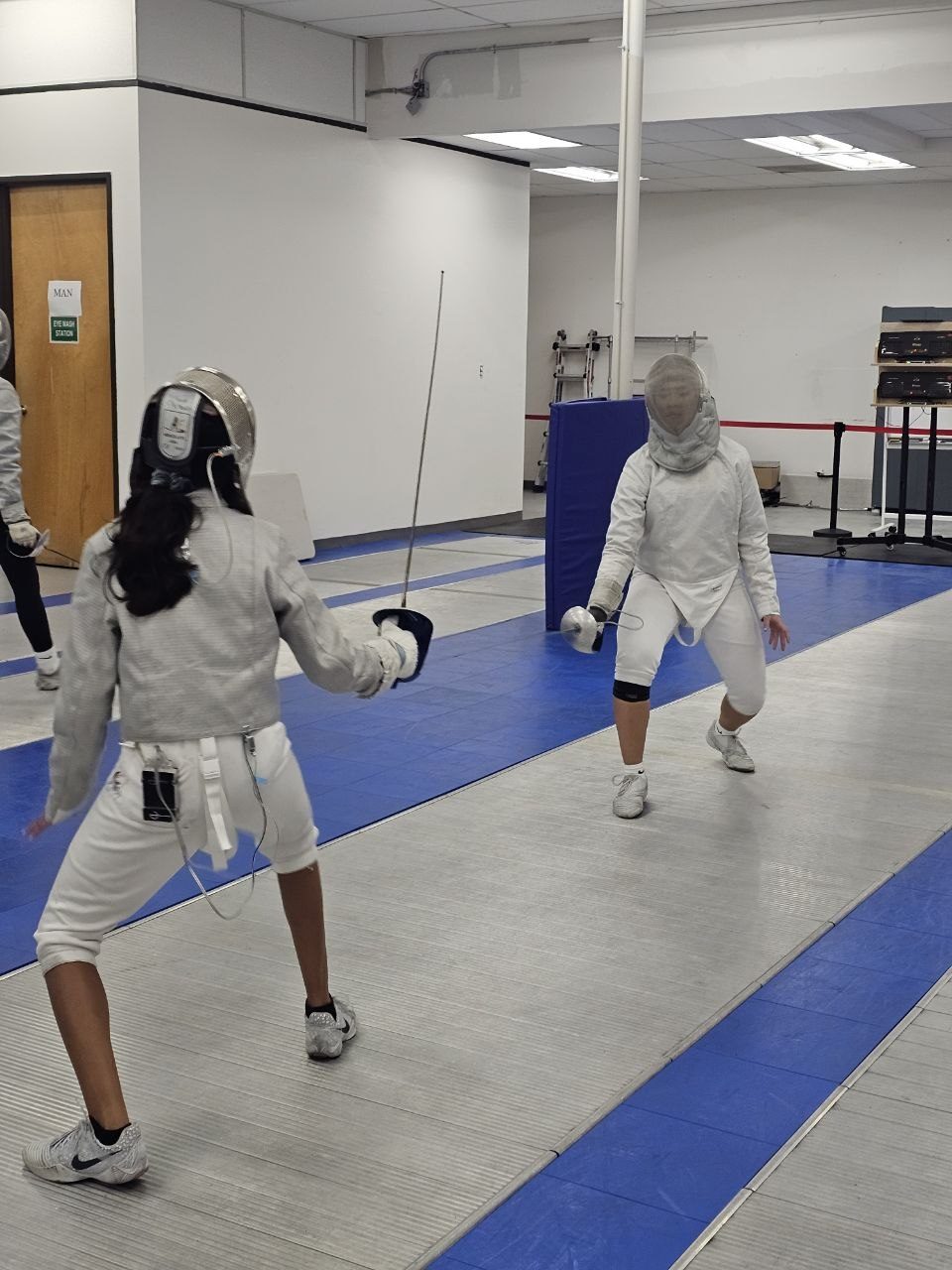 Two fencers in masks and white kit facing each other on the strip at West Coast Fencing Academy