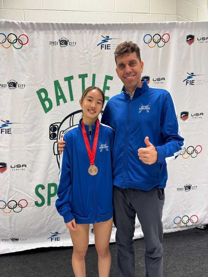A girl in a blue club jacket with a medal stands beside the coach, who gives a thumbs-up, in front of a tournament banner