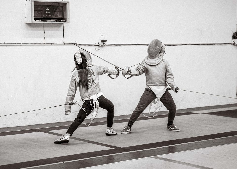 Black-and-white photo of two young fencers in masks and jackets on the strip, blades crossed, the one on the left in a deep lunge