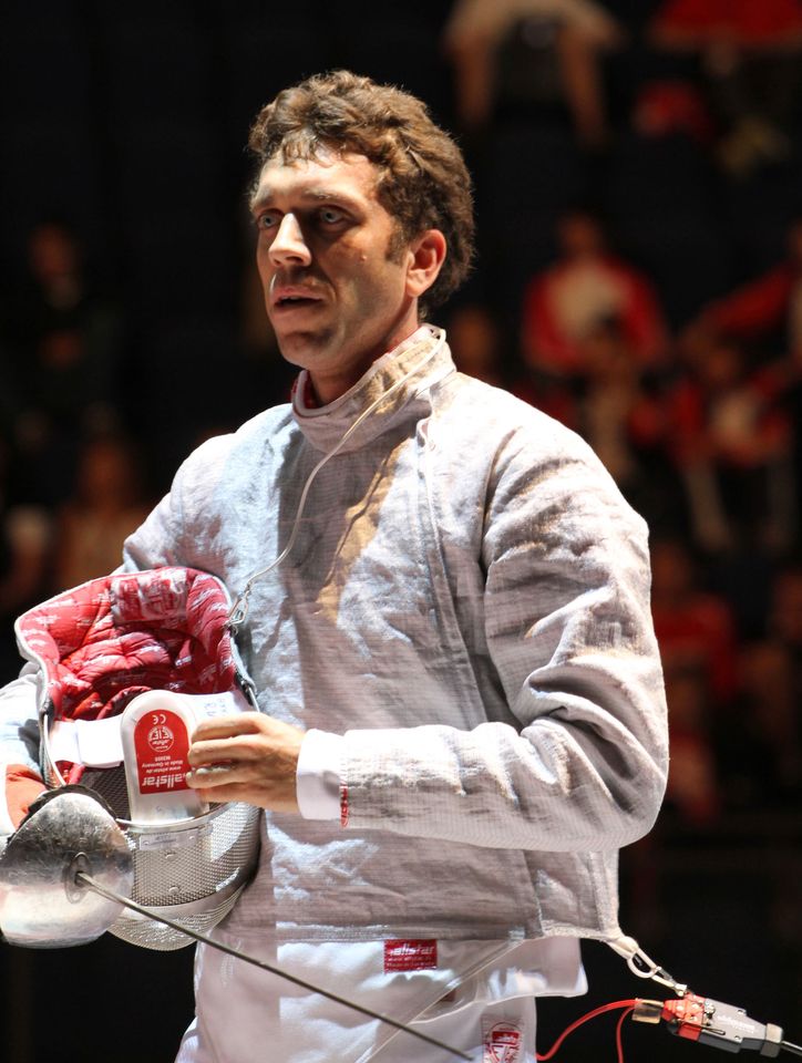 Pavel Bykov in a silver sabre lamé, body cord on, holds his mask under his arm in a dark arena