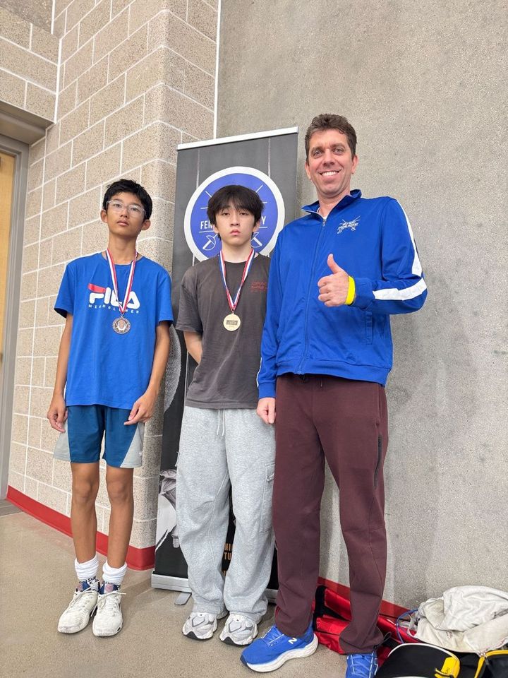Two boys with medals stand beside the coach, who gives a thumbs-up, in front of a West Coast Fencing Academy banner