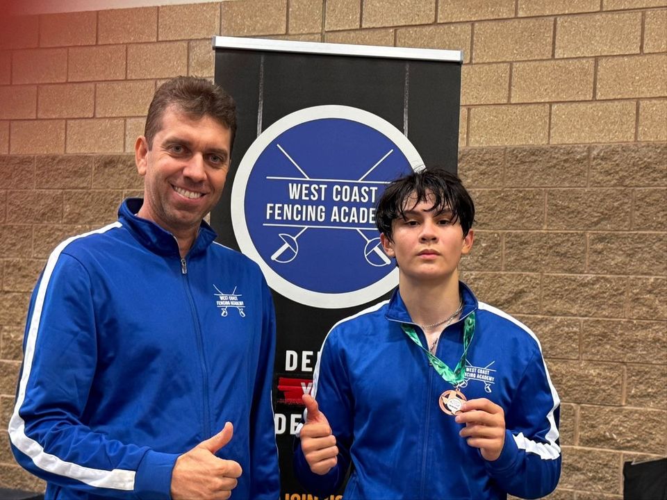 A coach and a teenage fencer in matching blue club tracksuits; the fencer holds up a medal and the coach gives a thumbs-up