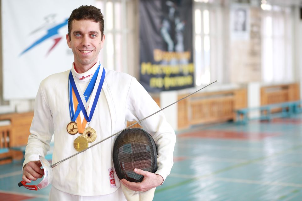 Pavel Bykov in fencing whites with his medals, holding his mask and sabre