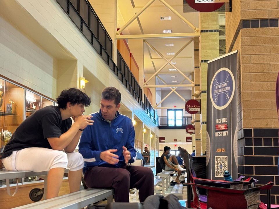The coach, in a blue club jacket, talks with a teenage fencer on a bench, a West Coast Fencing Academy banner behind them