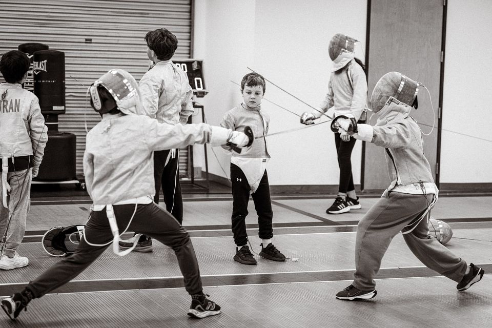 Black-and-white photo of children in fencing jackets on the strips: two in masks lunge with their sabres, a boy without a mask stands between them and others look on