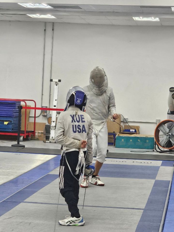 A young sabre fencer, back to the camera, a name and USA printed on the jacket, faces a taller masked fencer in white across the strip