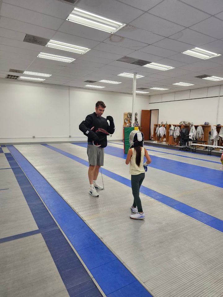 A coach and a young fencer work across a strip in the wide club salle
