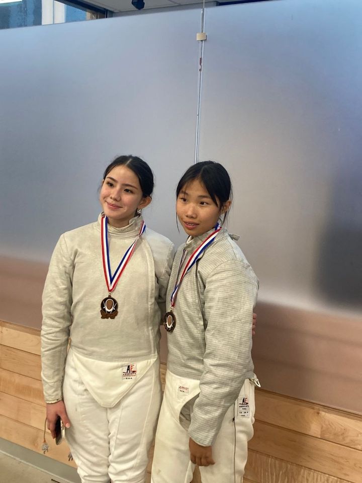 Two girls in white fencing jackets wear medals on red, white and blue ribbons