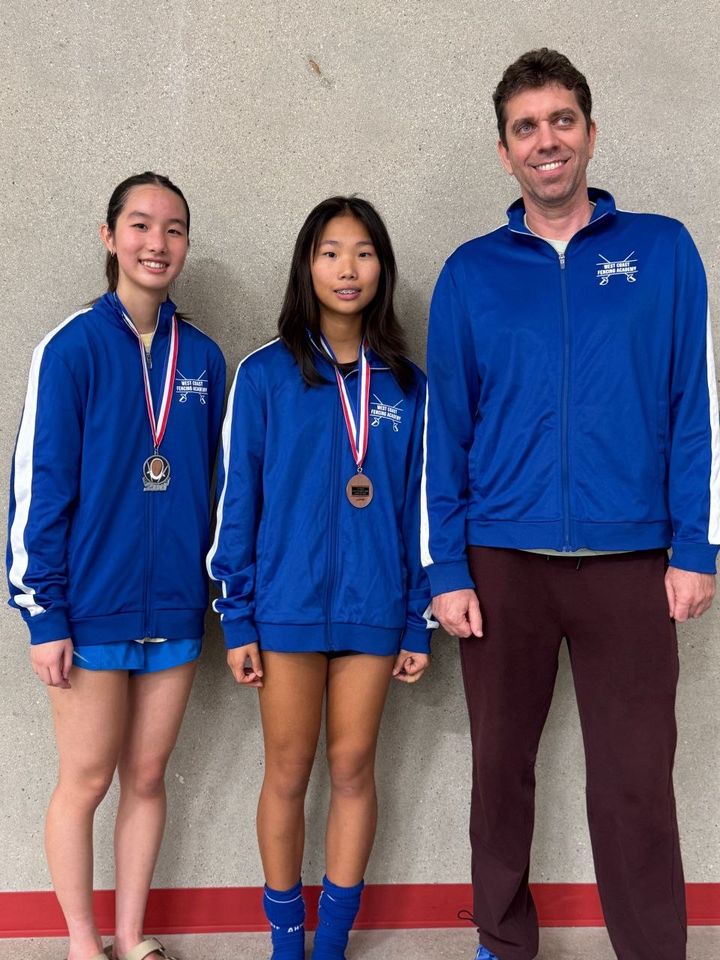 Two girls in blue club jackets wear medals and stand beside the coach against a gym wall