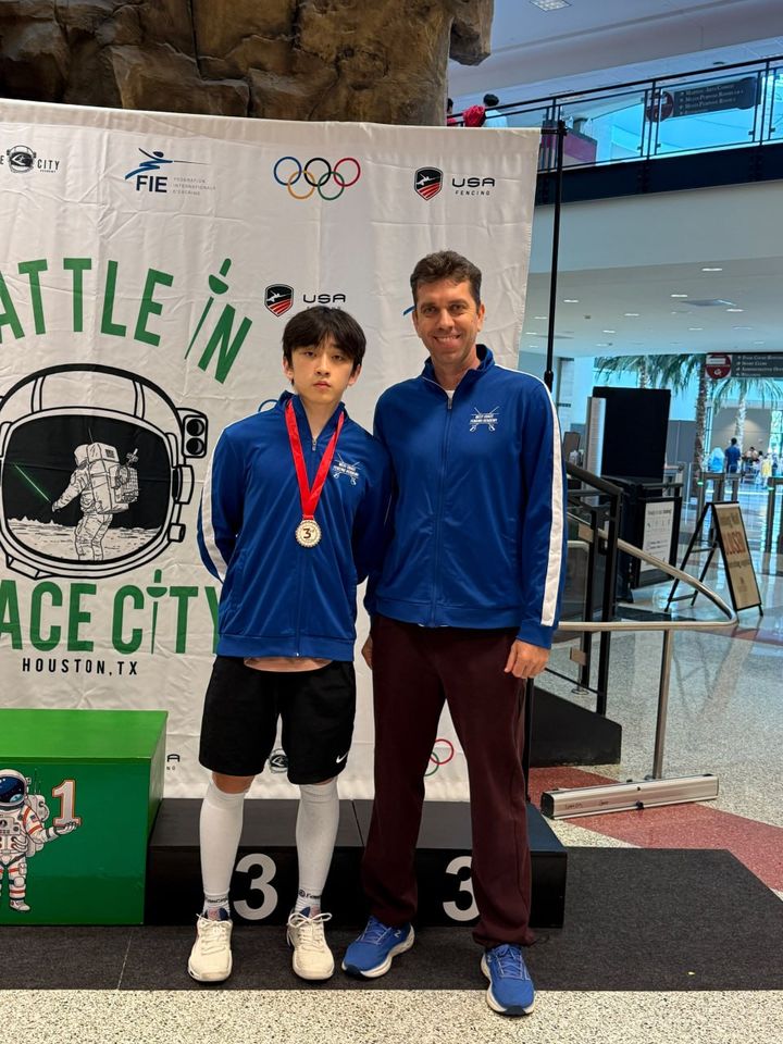 A teenage fencer with a medal stands by the podium beside the coach, in front of the “Battle in Space City, Houston, TX” banner