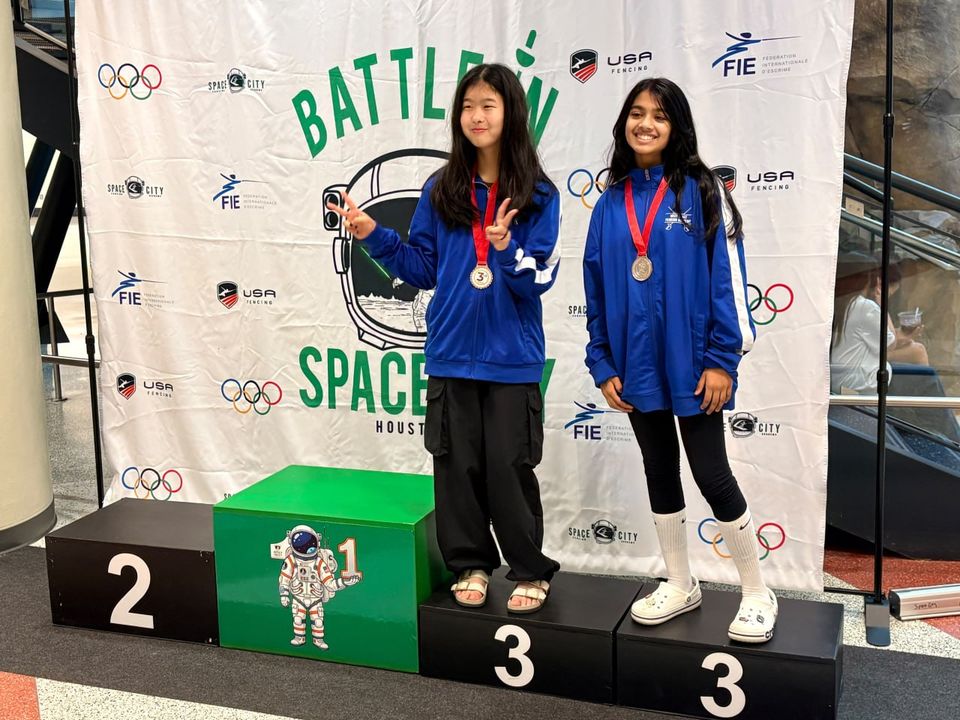 Two girls in blue club jackets, medals on, stand side by side on the podium; one makes a peace sign