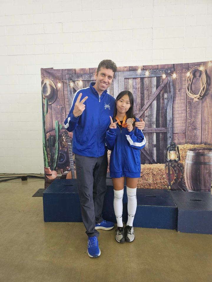 The coach and a young girl with a medal make peace signs by the podium, in front of a western-themed backdrop