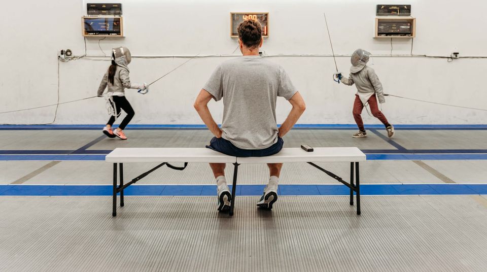 A man in a grey T-shirt sits on a bench, hands on his hips, and watches two young masked fencers on the strips, scoring machines on the wall above them