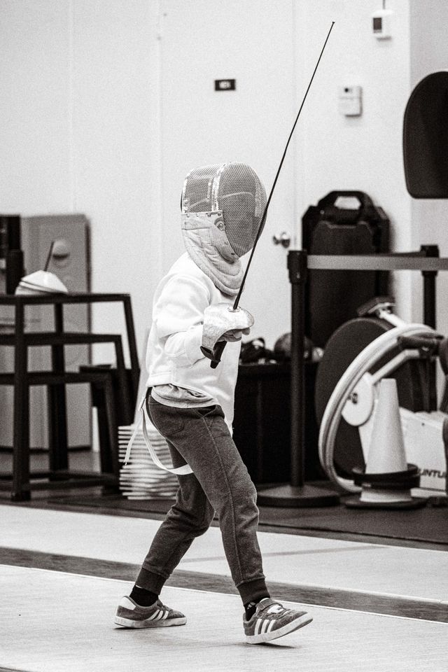 A young fencer in a mask and white jacket steps along the strip with the blade raised