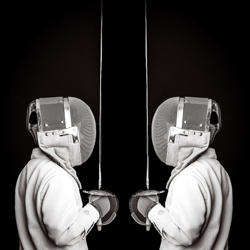 Two masked fencers in white jackets stand face to face on a black background, blades held upright in front of their masks