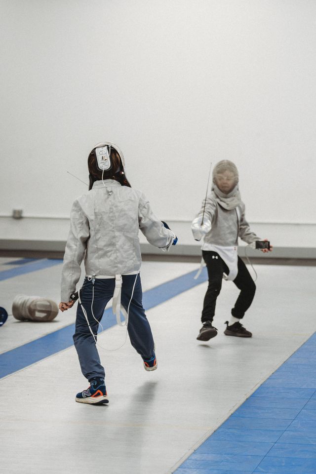Two young fencers in grey jackets face each other across a strip with blue edges; the nearer one, seen from behind, is wired to the body cord