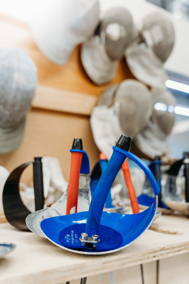 A blue sabre guard and grip resting on a wooden shelf, masks hanging out of focus behind it