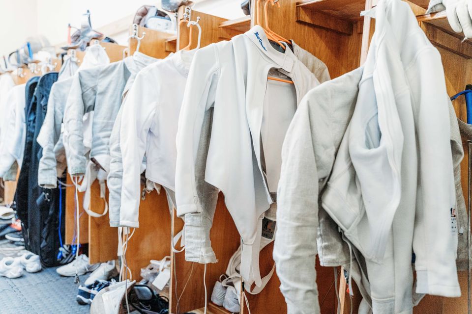 White and grey fencing jackets on hangers along a row of wooden cubbies, shoes on the floor below