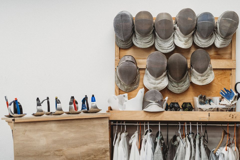 Fencing masks on a wooden rack, sabre guards lined up on a shelf beside it and white jackets on hangers below