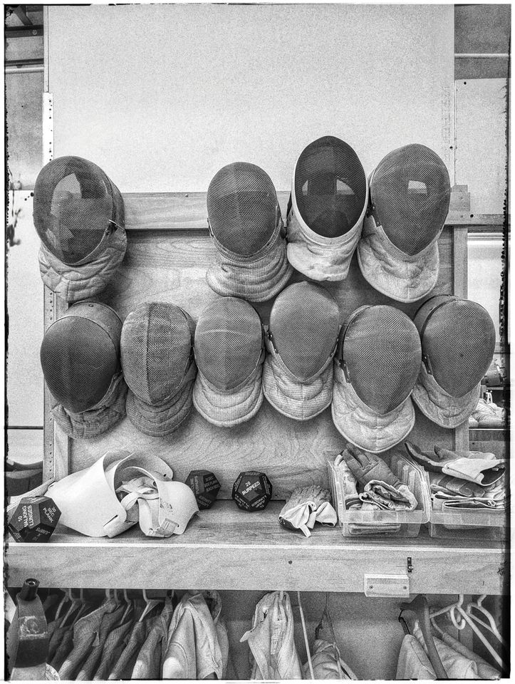 Black-and-white photo of fencing masks hanging in two rows on a wooden rack, gloves and exercise dice on the shelf below