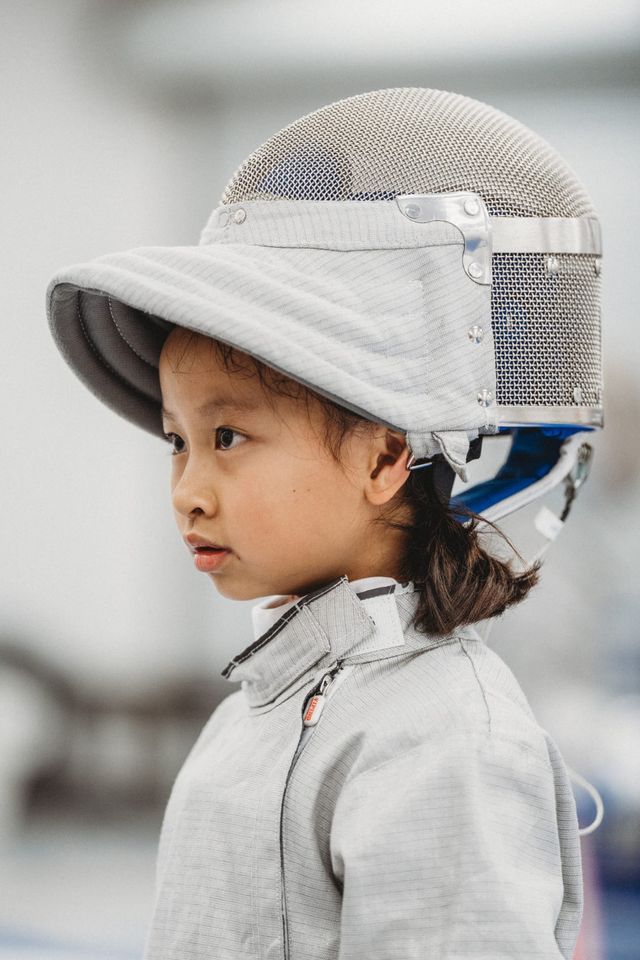 A young girl in a grey fencing jacket, her mask pushed up on top of her head, looks across the room
