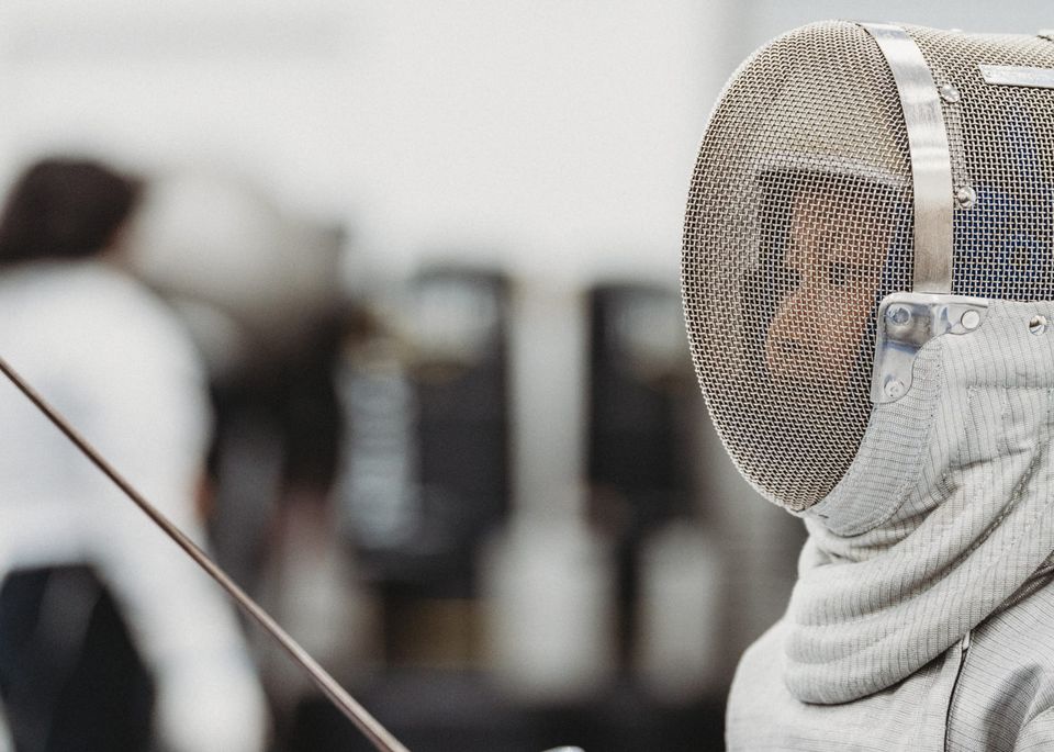 A close-up of a fencer in a mesh mask and padded bib, the face just visible through the mesh