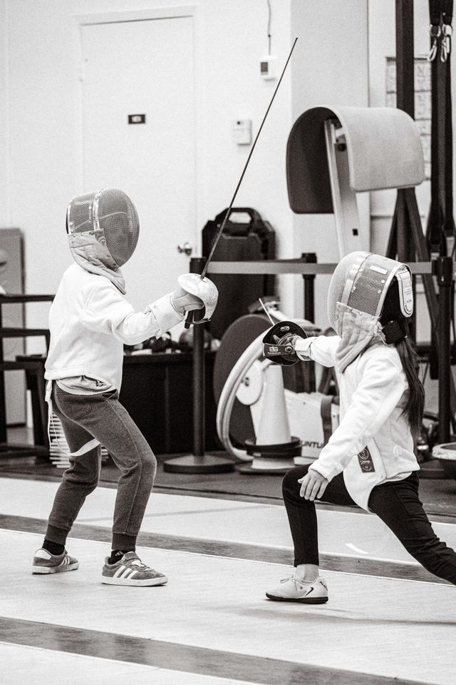 Two young fencers in white jackets and masks on the strip: one holds the blade high, the other lunges in under it