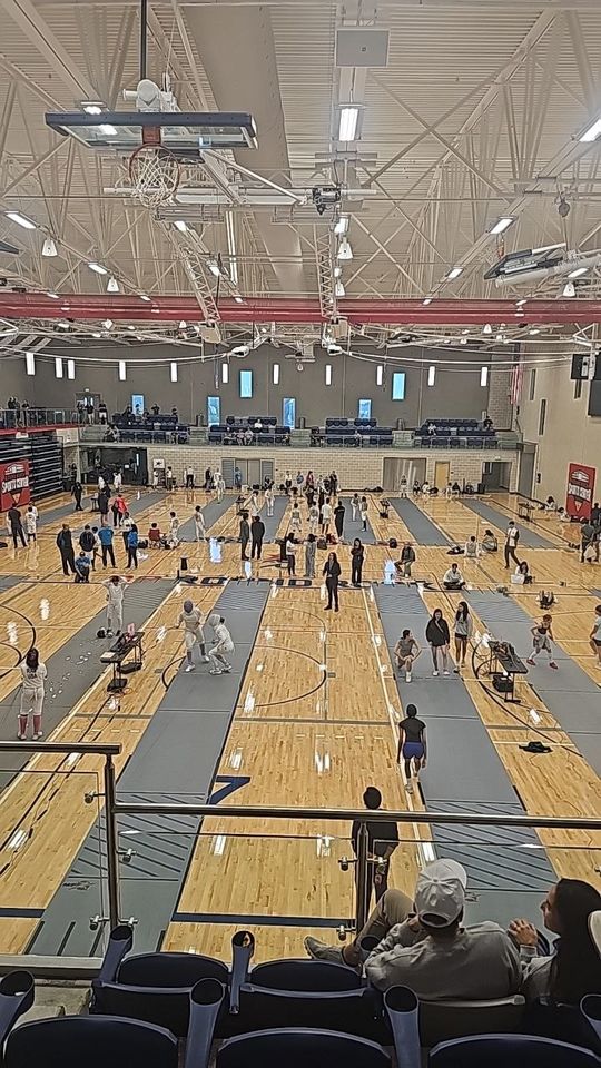 A large sports hall seen from the stands, its floor laid with rows of fencing strips and fencers on them