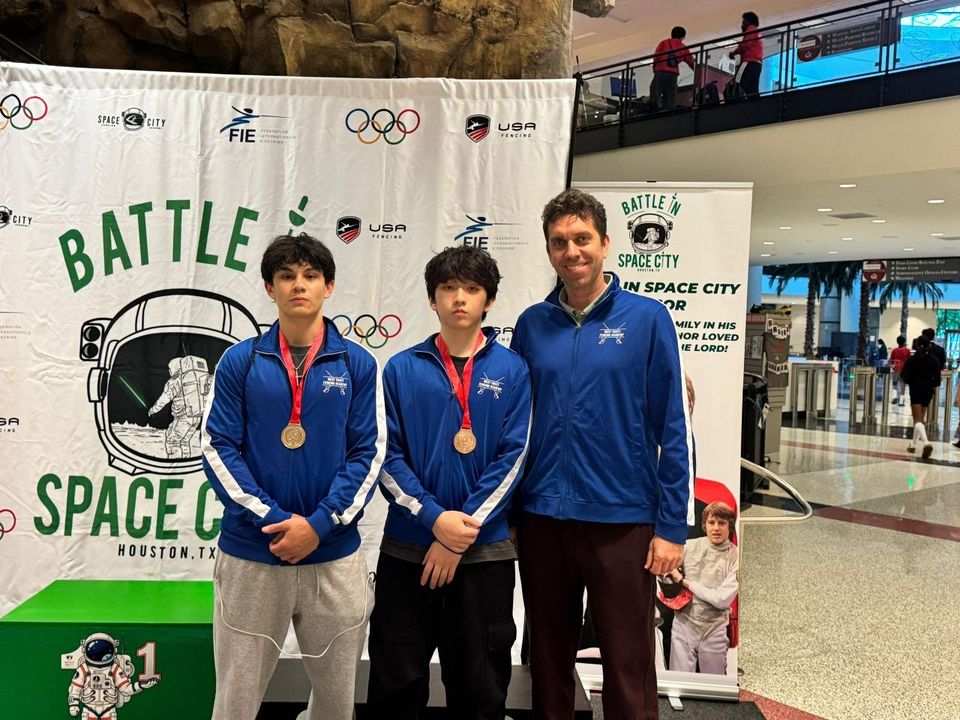 A coach and two teenage fencers in blue West Coast Fencing Academy tracksuits, medals on, in front of a “Battle in Space City, Houston, TX” banner