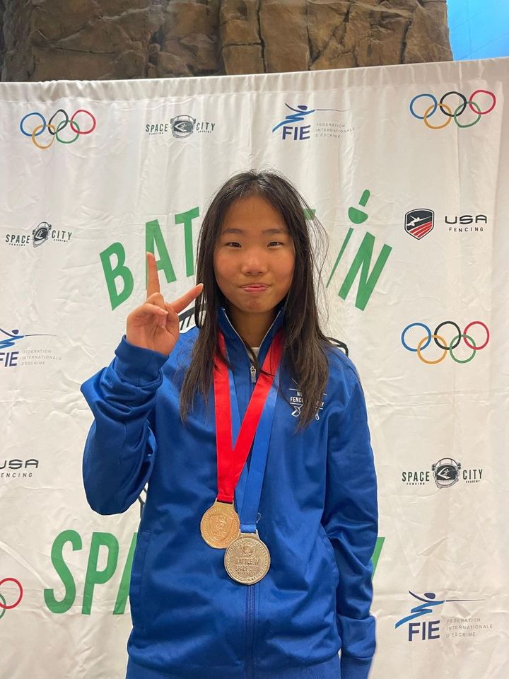 A girl in a blue club jacket wears two medals and makes a peace sign in front of a tournament banner