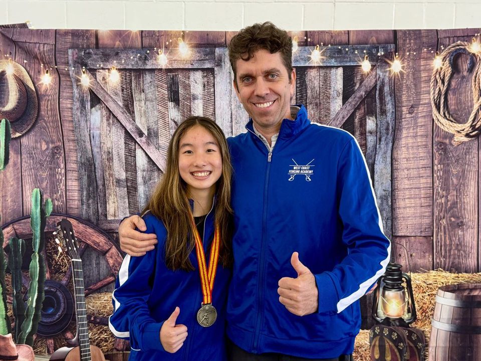 A teenage girl with a medal and the coach both give a thumbs-up in front of a western-themed backdrop